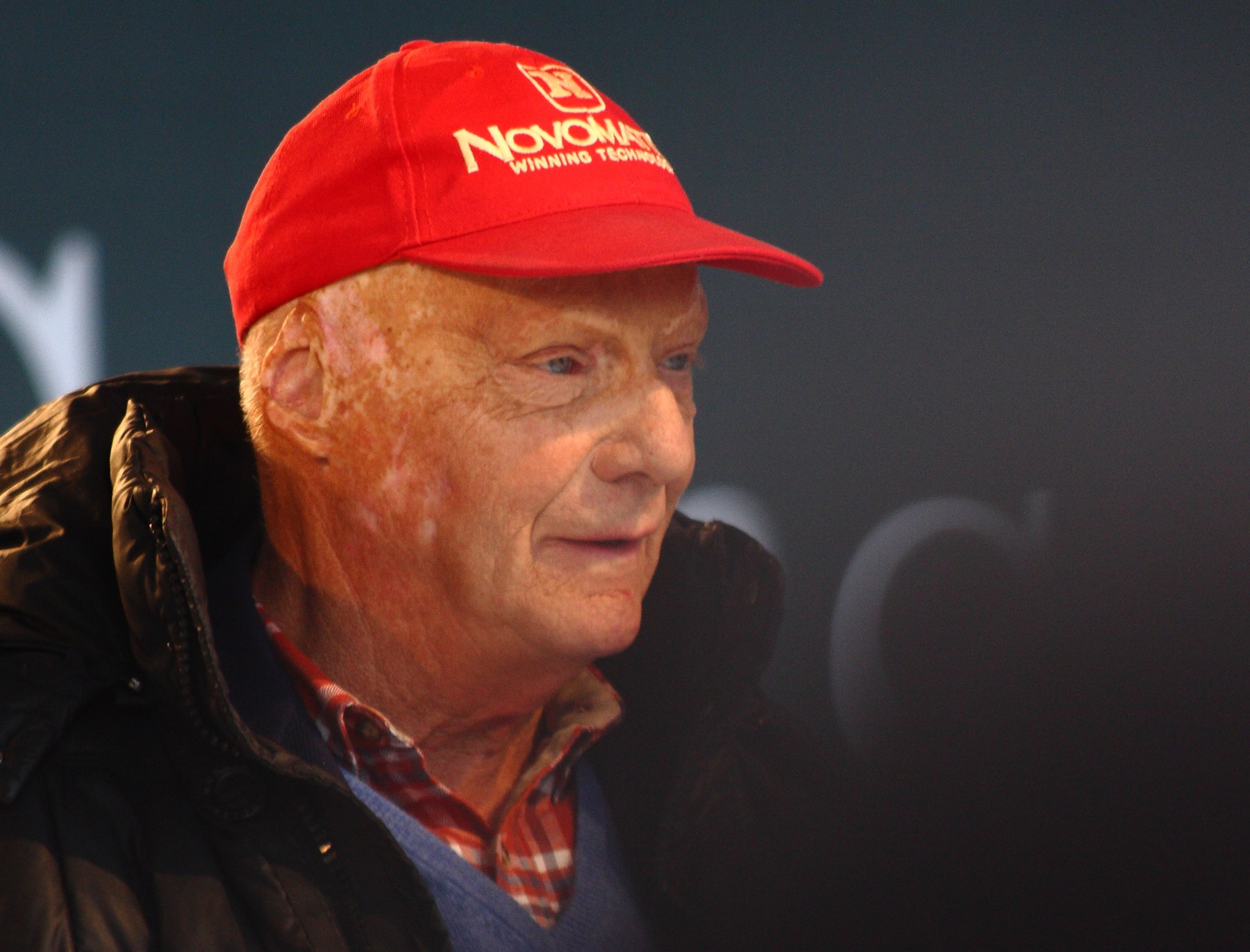In colour: Lauda in profile, smiling, in a red cap and a dark padded jacket over a checked shirt and pullover, against a dark stage backdrop.