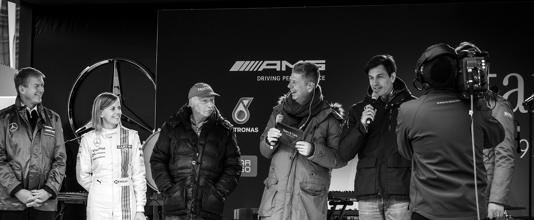 A line of people on an outdoor stage before a large three-pointed-star emblem and sponsor boards: a smiling man, a woman in a striped race suit, Lauda in a cap and padded jacket, a presenter and a man speaking into a microphone, with a television cameraman at right.