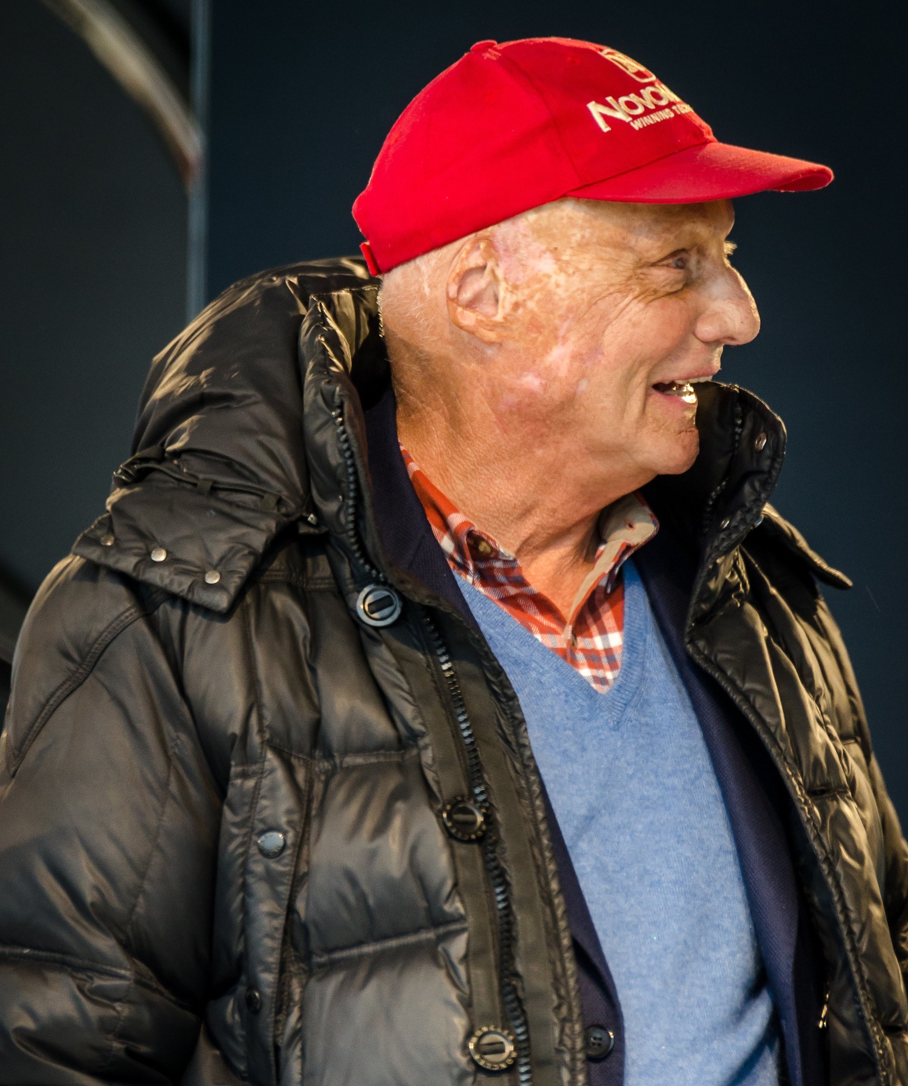 In colour: Lauda, seen from the side, laughing, in a red sponsor’s cap, a dark padded jacket, a checked shirt and a pale blue pullover, against a dark backdrop.
