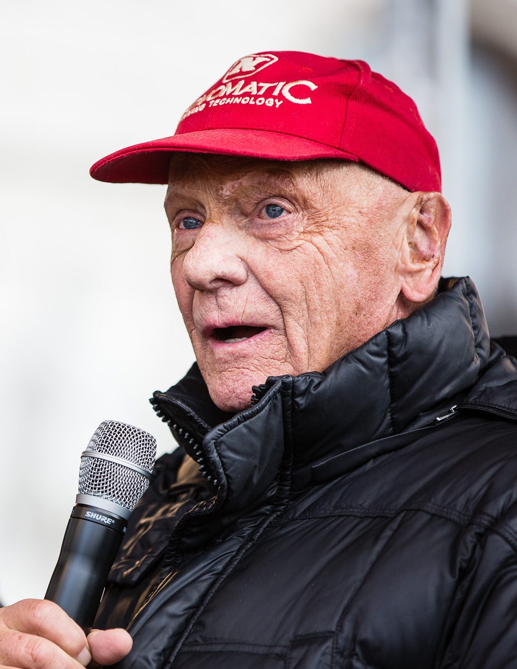 In colour: Lauda in mid-sentence, in a red cap and a black padded jacket, holding a microphone, against a pale background out of focus.