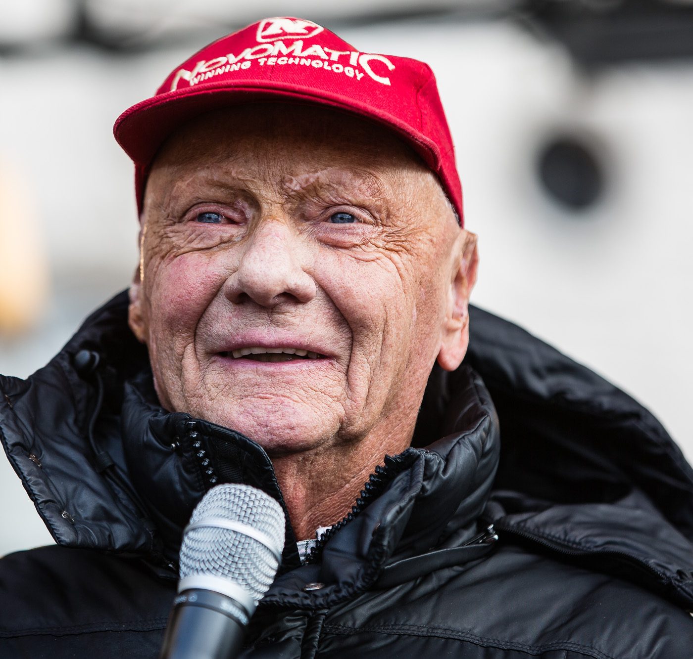 In colour: Lauda, smiling, in a red cap and a black padded jacket, a microphone in front of him, against a soft pale background.