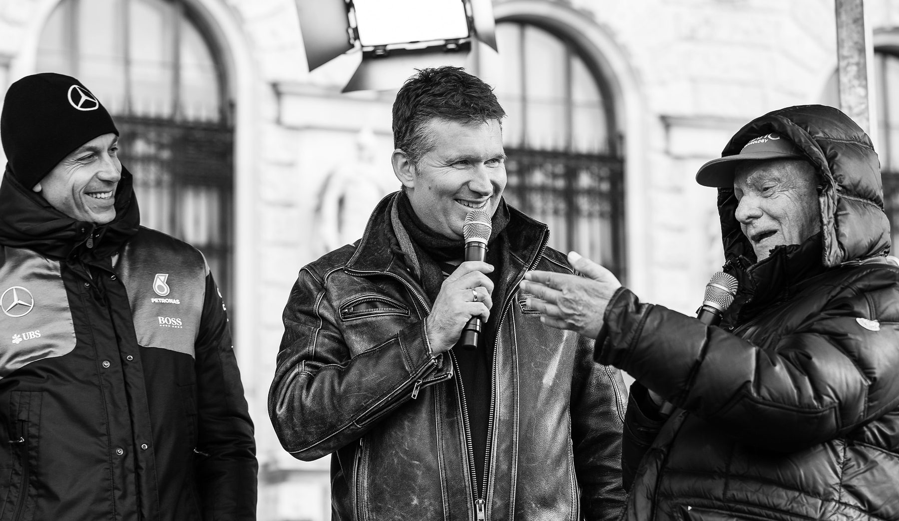 Three men on an outdoor stage before a stone façade with arched windows: at left a laughing man in a team beanie and jacket, in the middle a presenter in a leather jacket with a microphone, at right Lauda in a hood and cap, gesturing as he speaks.