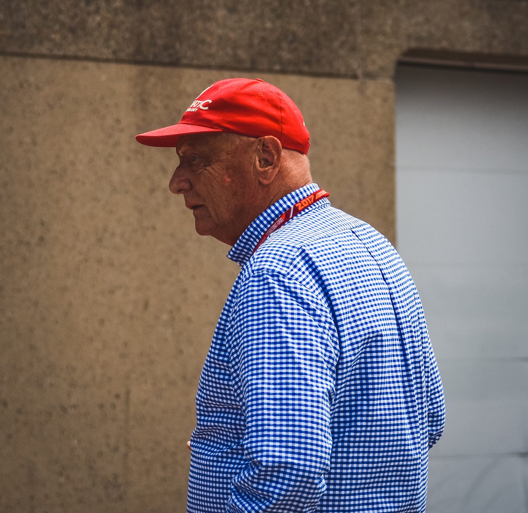 In colour: Lauda in profile, in a red cap and a blue gingham shirt, walking past a garage wall.
