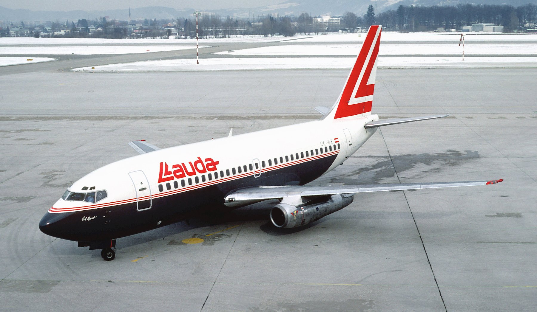 In colour: a Boeing 737 in white with a dark belly, red “Lauda” titles and a large red L on the fin, stands on a grey apron. Snow lies on the grass beyond, with a town and hazy hills behind.