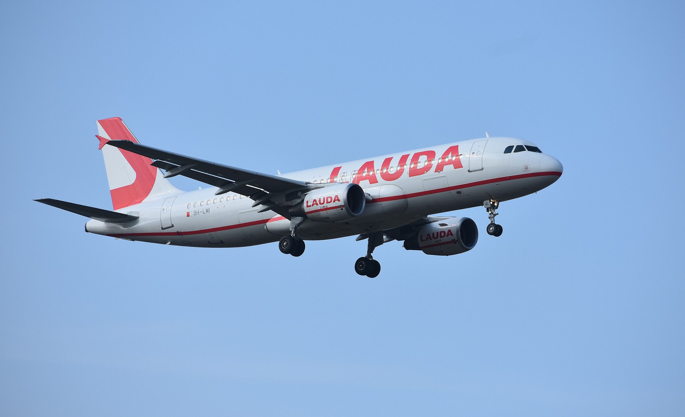 In colour: a white Airbus A320 with LAUDA in large red letters along the fuselage and on both engines, and a red tail, flies with its landing gear down against a clear blue sky.