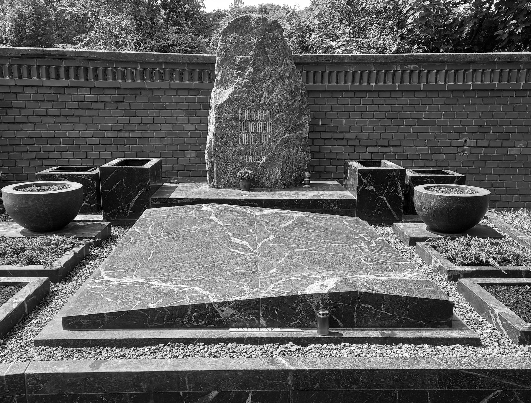 A family grave: a tall rough-hewn dark stone carved with a short inscription and the family name, a polished marble slab, two stone bowls and grave lights, before a brick wall and shrubs.