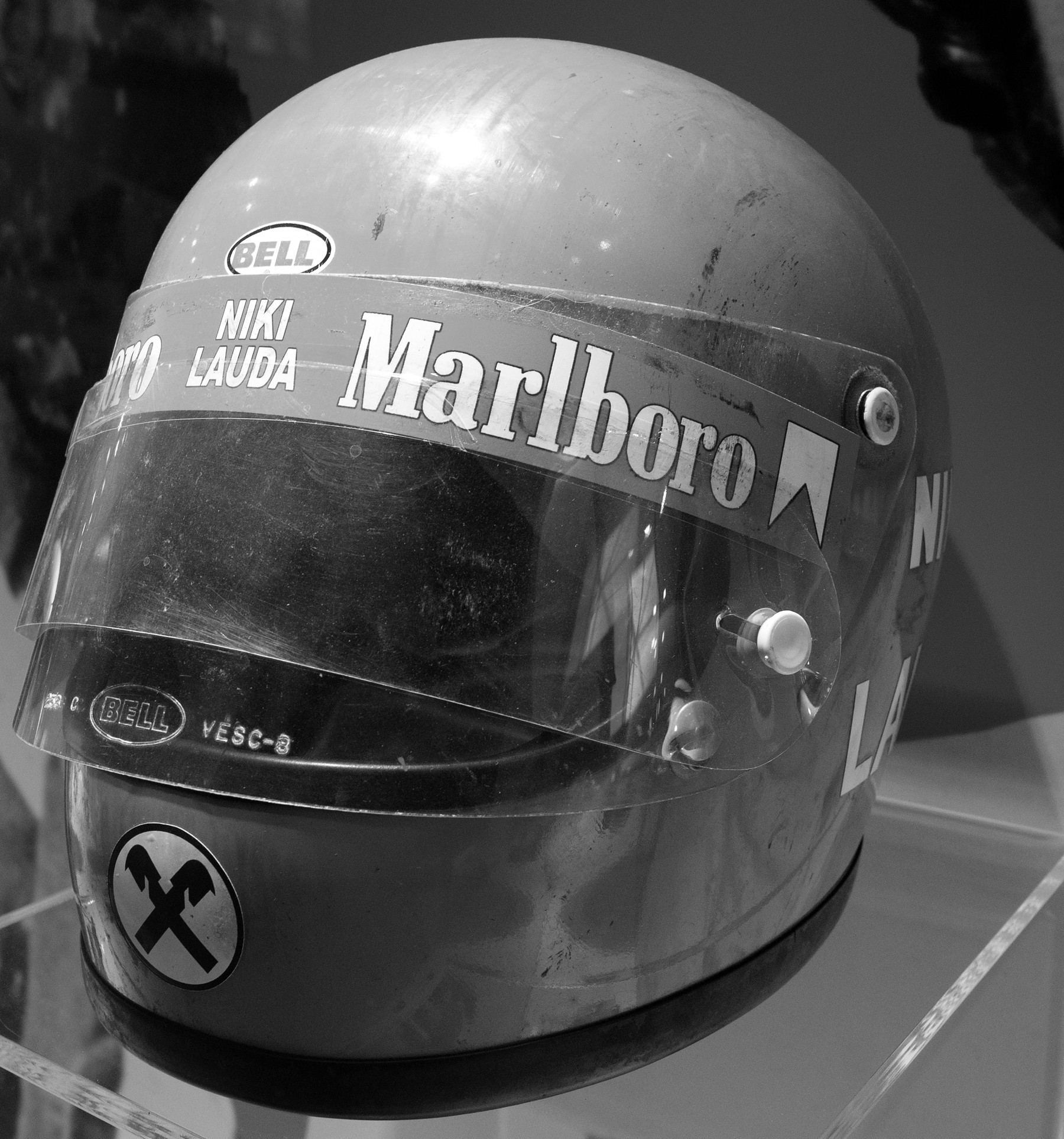 A worn, scuffed full-face racing helmet on a clear stand in a museum display, a sponsor’s band with the name NIKI LAUDA across the top of the visor and a round emblem of two crossed gables on the chin.