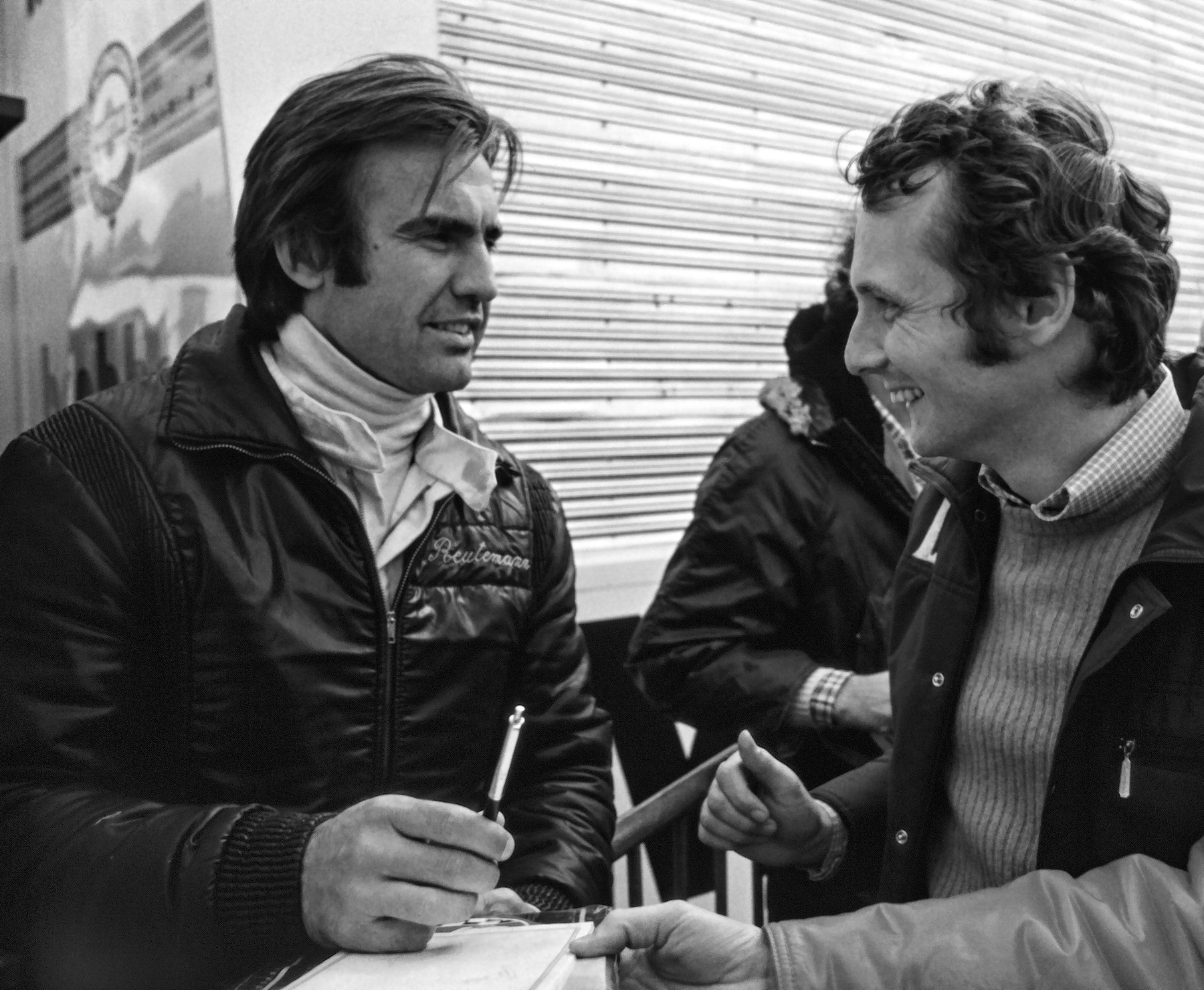 Two men in padded jackets in a paddock: the one at left, his name stitched on his chest, holds a pen over a paper; the one at right, curly-haired, grins at him. A corrugated trailer wall behind, a third man between them, and a fan’s arm across the bottom right.