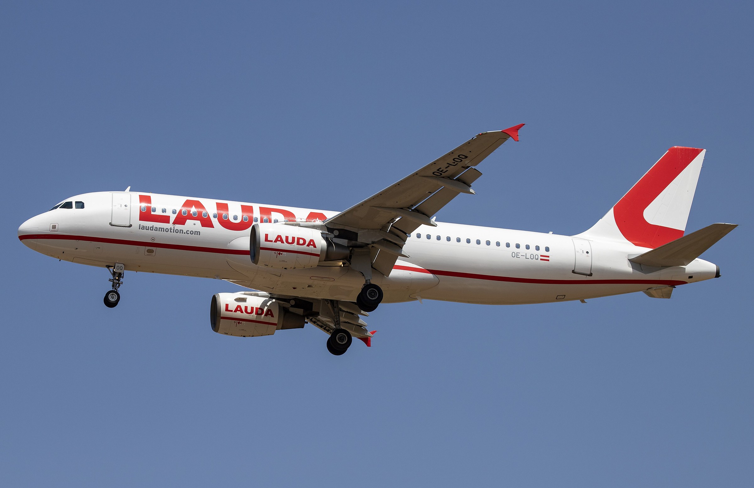 In colour: a white Airbus A320 with “LAUDA” in large red letters along the fuselage and on both engines, a red tail and red wingtips, flies overhead with its landing gear down against a deep blue sky.