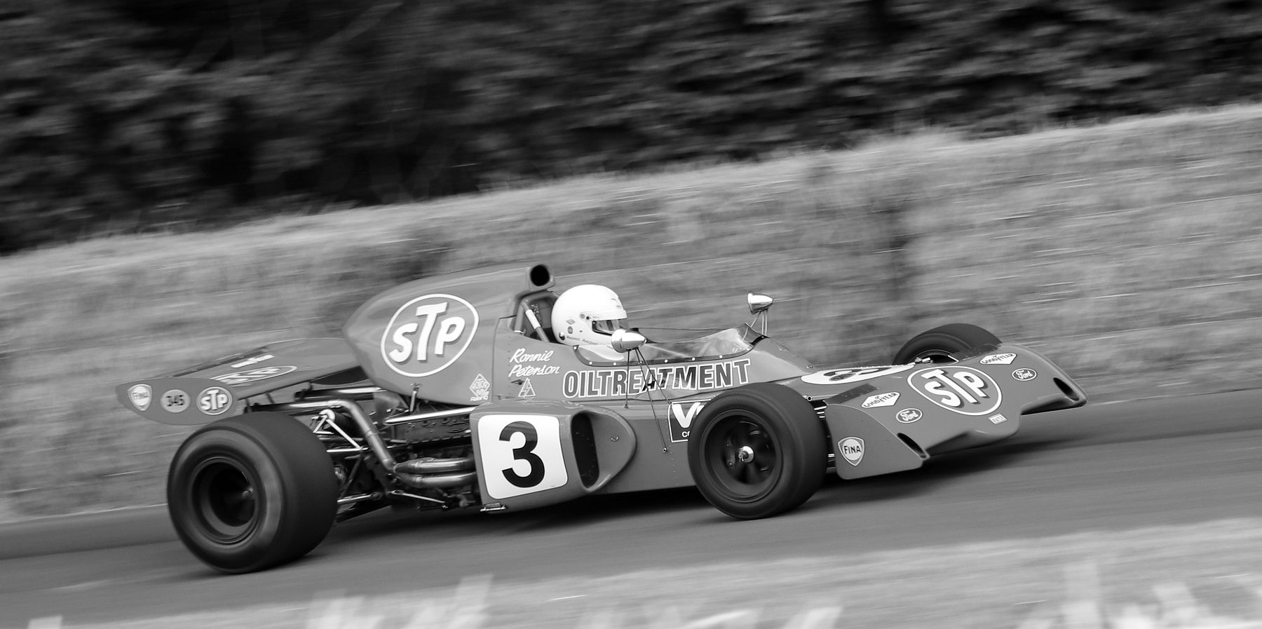 A 1970s single-seater with a tall airbox, No. 3 on its side and the name Ronnie Peterson on the cockpit, at speed past straw bales, the trees behind streaked by the panning camera.