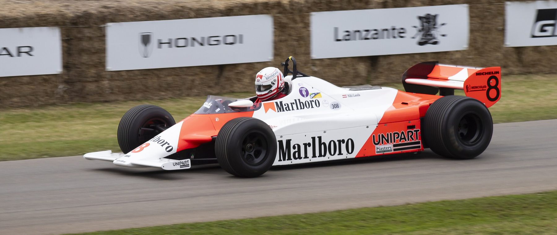 In colour: a red and white Formula One car with No. 8 on its rear wing, at speed past straw bales and advertising boards, its driver in a white helmet.