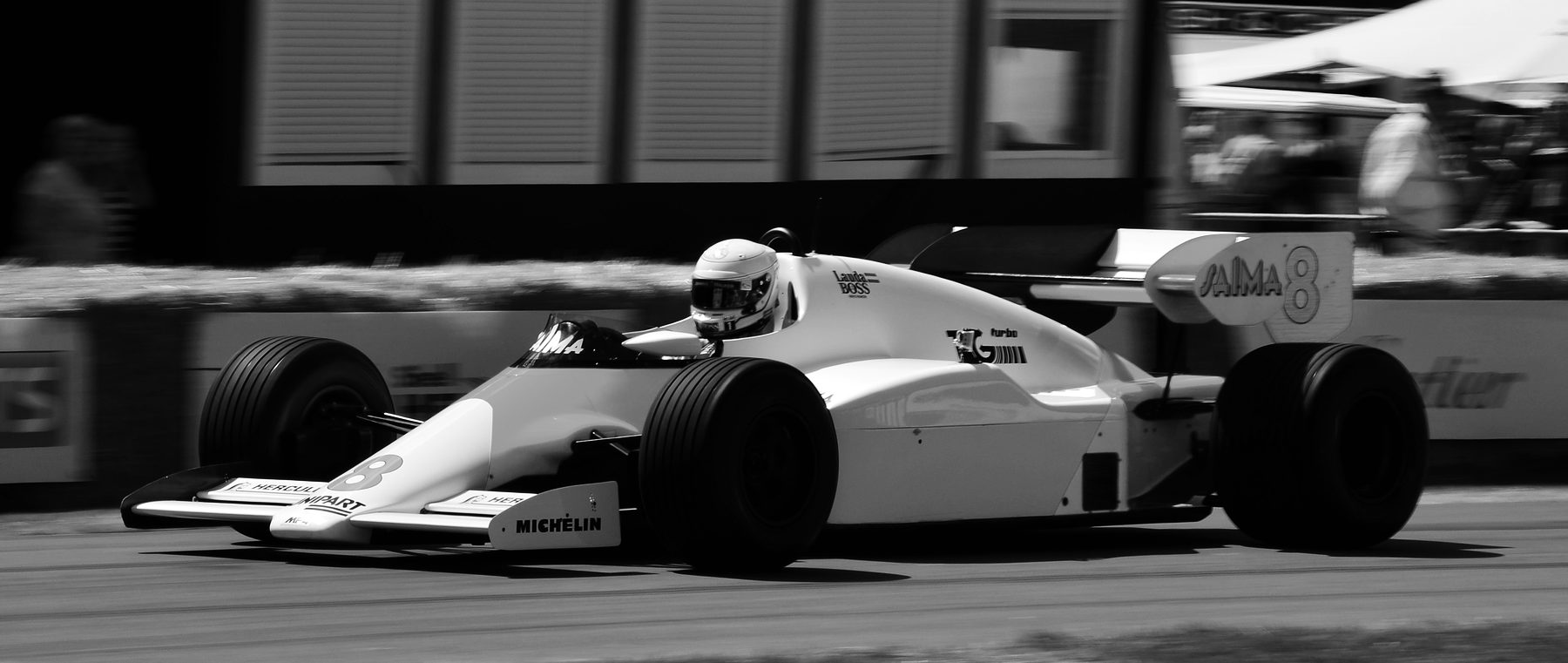 A two-tone turbo-era Formula One car with No. 8 on its nose and rear wing, its driver in a pale helmet, at speed in front of straw bales; spectators stand at the edges, blurred by the panning camera.