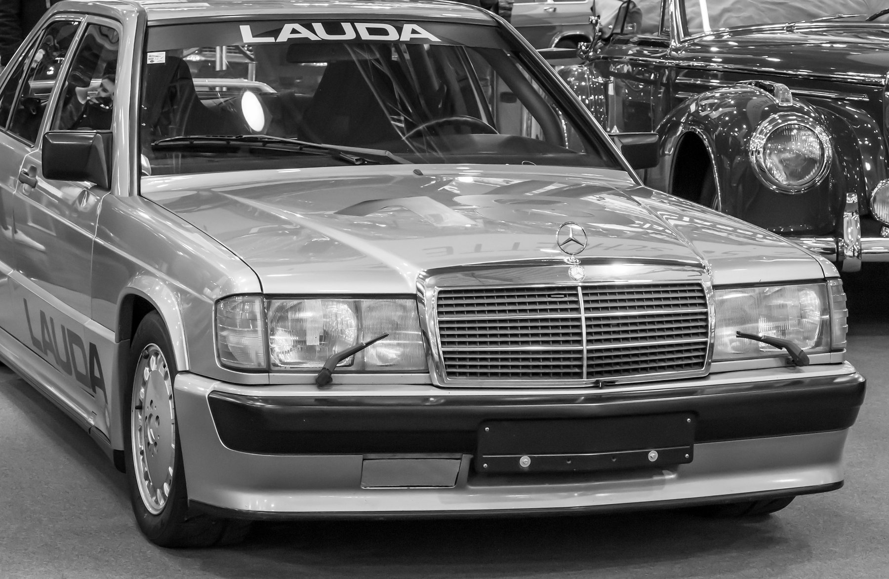 Front three-quarter view of a 1980s saloon with “LAUDA” across the top of the windscreen and along the doors, parked on a show-hall carpet beside a 1950s saloon.