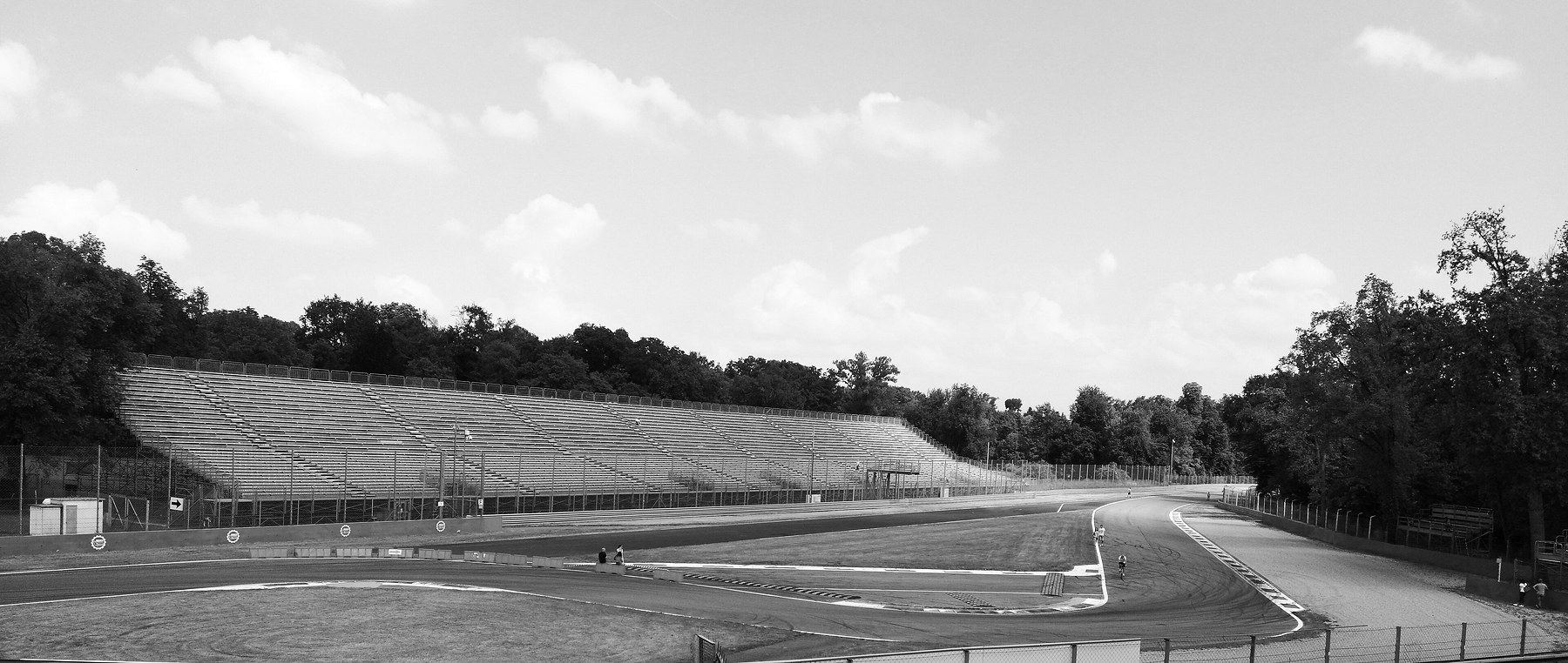 A chicane on a racing circuit seen from above the track: a tight left and right with striped kerbs and a row of plastic barriers, an empty tiered grandstand under trees behind, and a few people walking and cycling on the track.