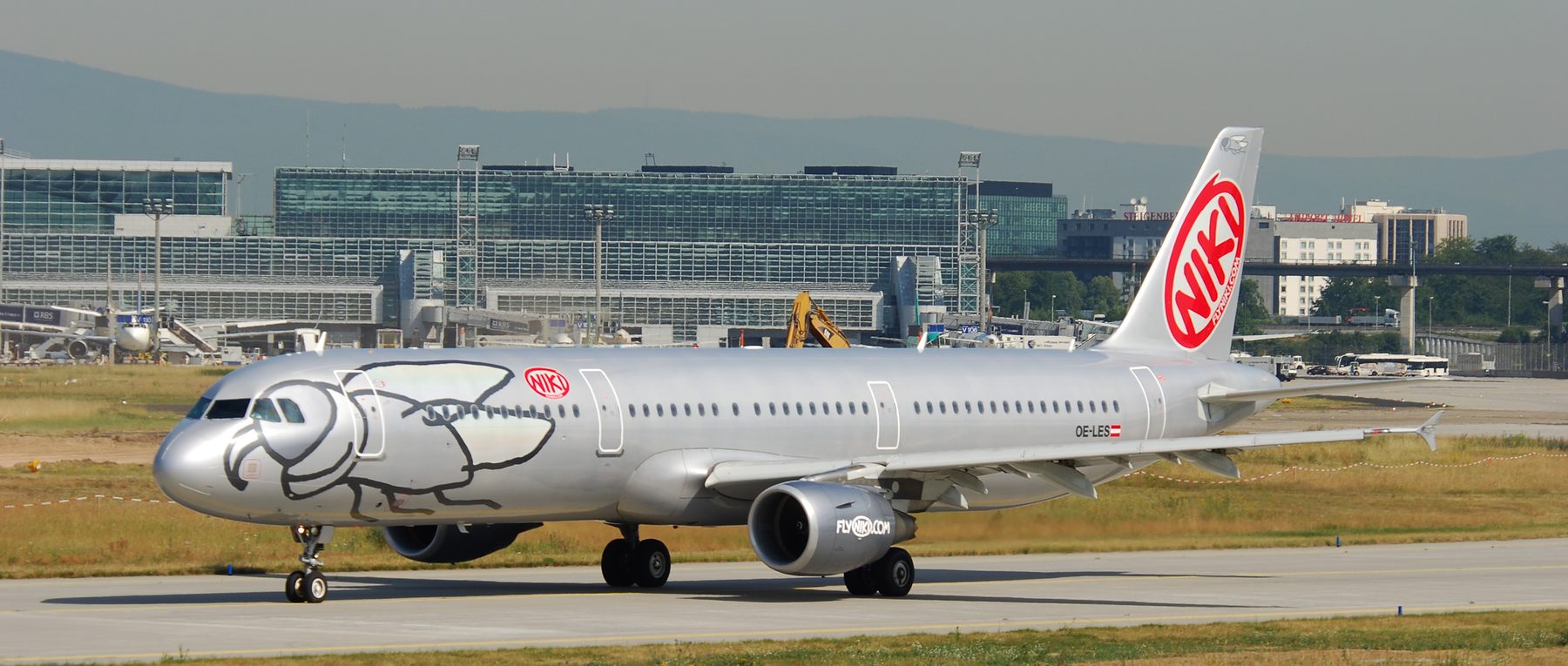 In colour: a silver Airbus A321 with a large black line drawing of a bird on its nose and a red “NIKI” disc on the tail taxies past the glass terminal buildings of Frankfurt airport, with dry summer grass in front and hazy hills behind.