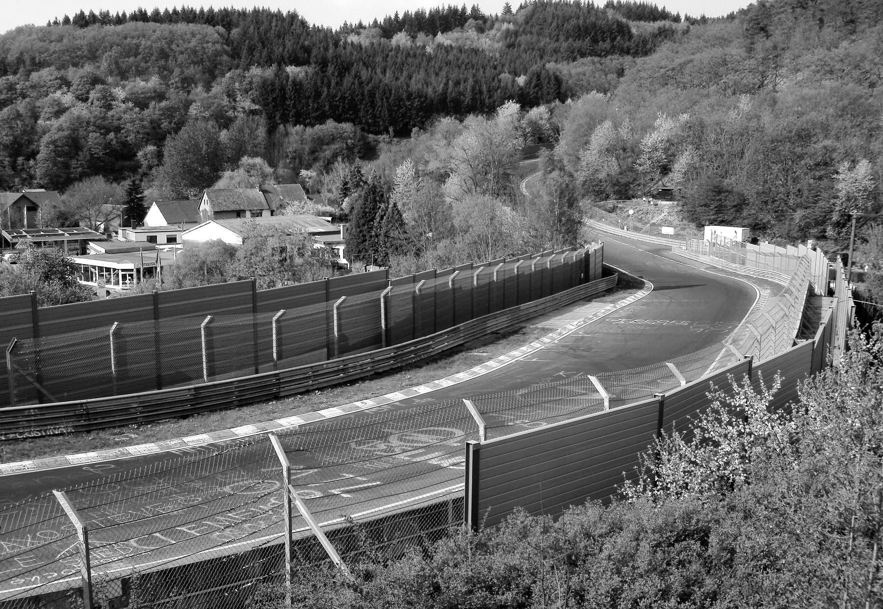 A racing circuit curving between tall fences and barriers through a wooded valley in spring, chalk writing on the track, striped kerbs, and houses and a filling station of a village beyond.