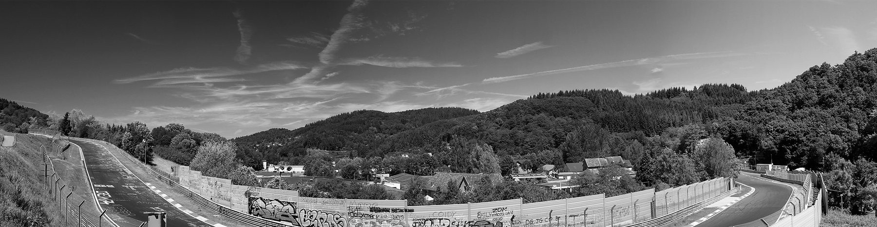 A wide panorama of a racing circuit curving round a village in a wooded valley under a clear sky, the track lined with striped kerbs and fences covered in graffiti, wooded hills rising behind.