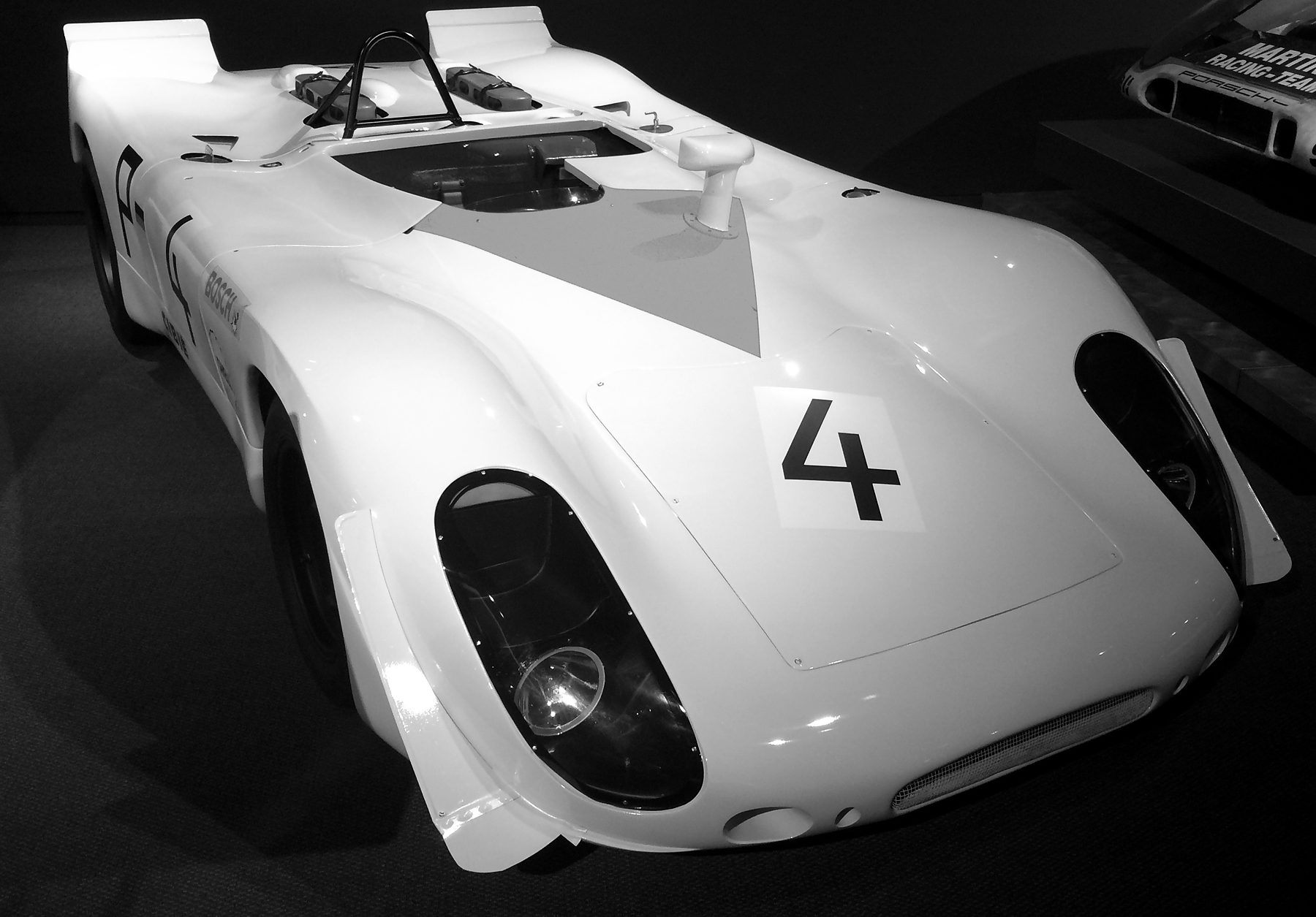 A low open sports-racing car with tall rear fins, a roll hoop and a large No. 4 on its nose, under spotlights on a dark floor; the nose of another racing car shows at the right edge.