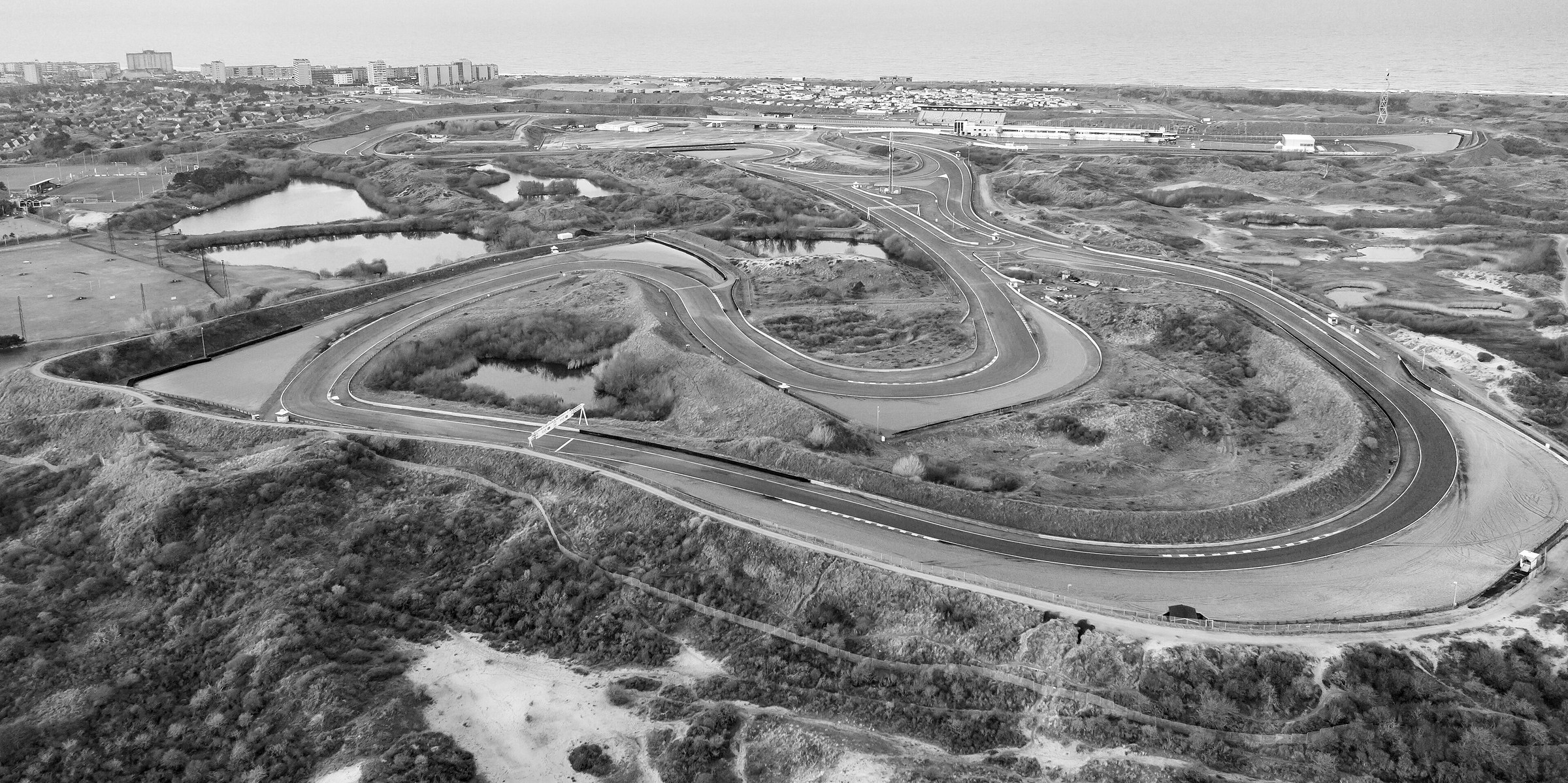 An aerial view of a racing circuit winding among sand dunes, scrub and small lakes, with pit buildings and a grandstand near the sea, tower blocks of a seaside town on the left and the sea on the horizon.